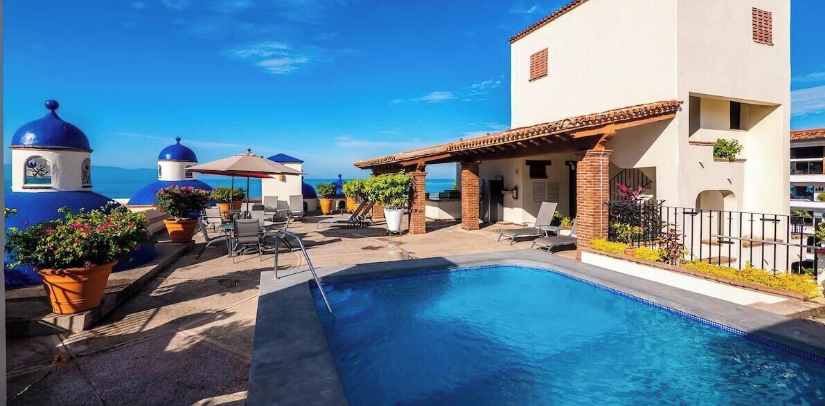 Un área de piscina con sillones, plantas en macetas y un edificio con cúpulas azules.