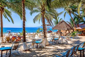 A beach with rows of sun loungers and palm trees, a thatched-roof hut, and a view of the ocean. - Ground-floor Condo With Resort Pool, Balcony, & Pool Tables (Nuevo Nayarit)