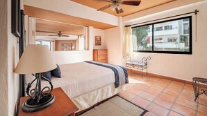 A bedroom with a bed, a nightstand, a lamp, a ceiling fan, a window with a view, and a small table with chairs. - One Beach Street 307 B· Amazing 2 BR Condo (Puerto Vallarta)