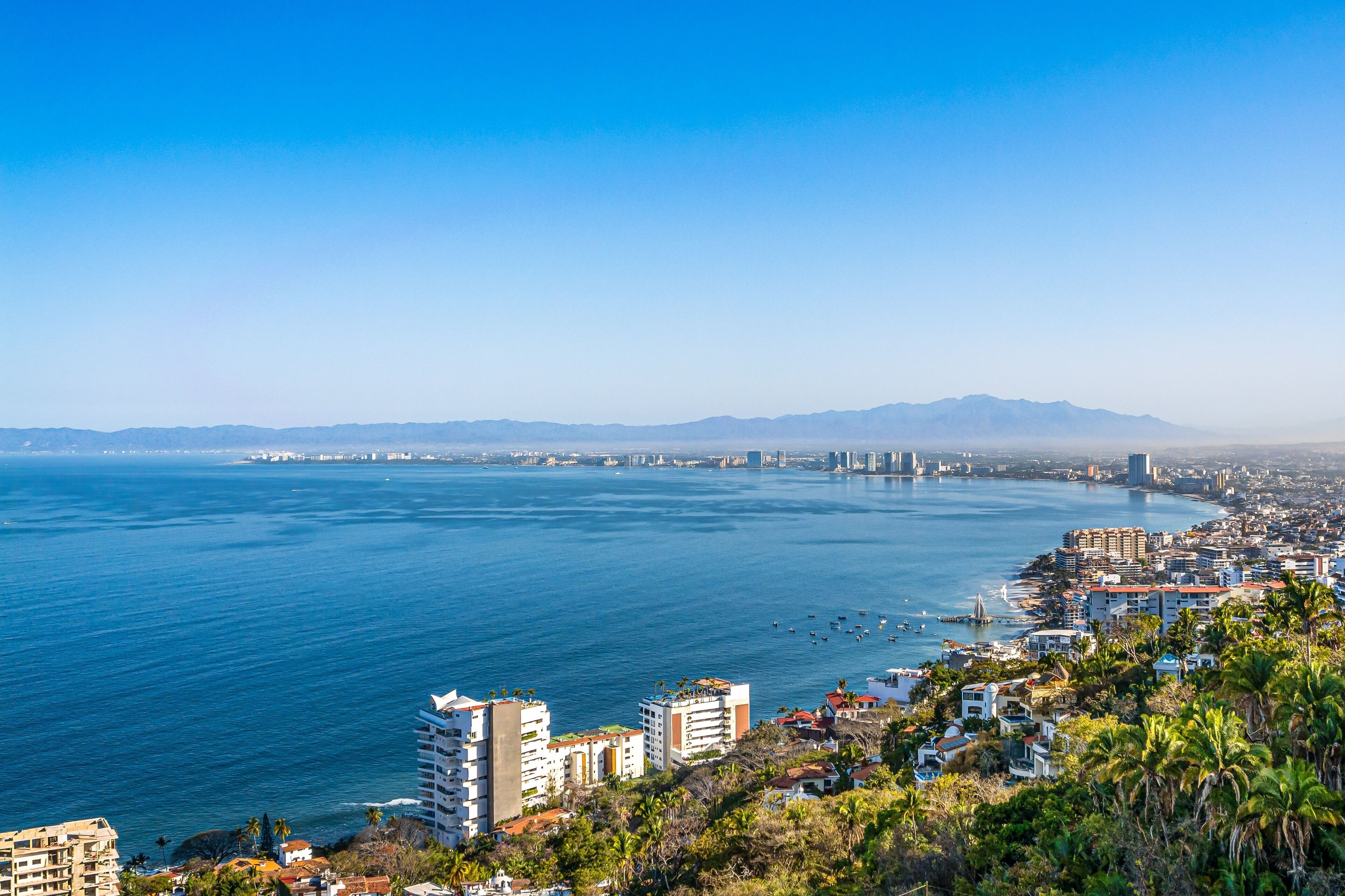 A coastal cityscape with a view of the sea, buildings, and mountains in the distance.