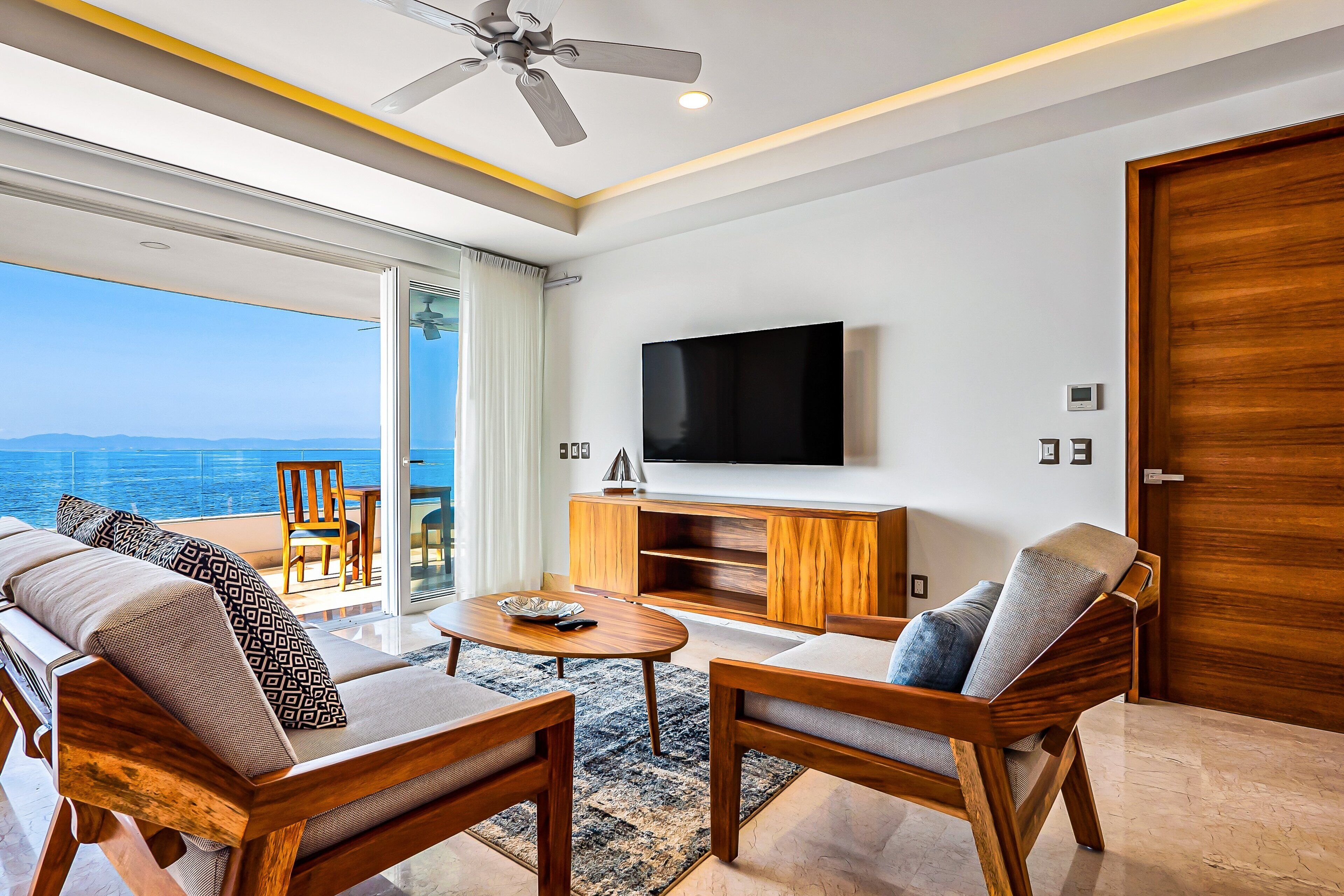 Un salón moderno con un televisor grande, muebles de madera y vista al mar.