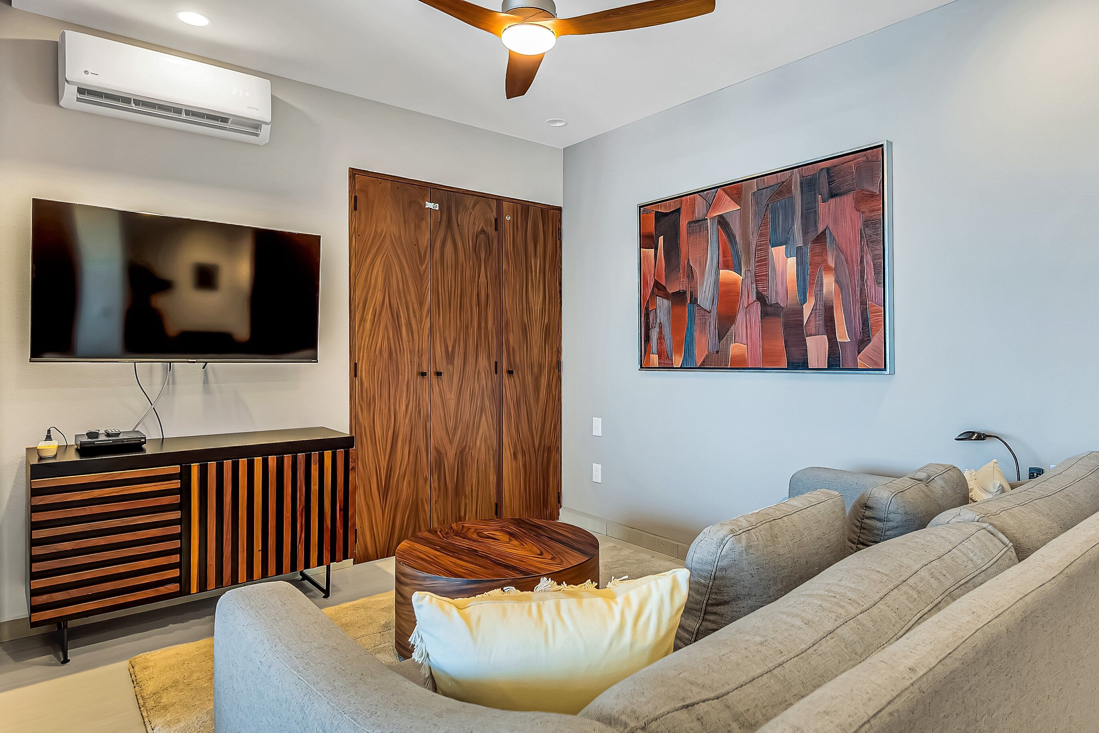 A modern living room with a flat-screen TV, wooden furniture, a ceiling fan, and a large abstract painting.