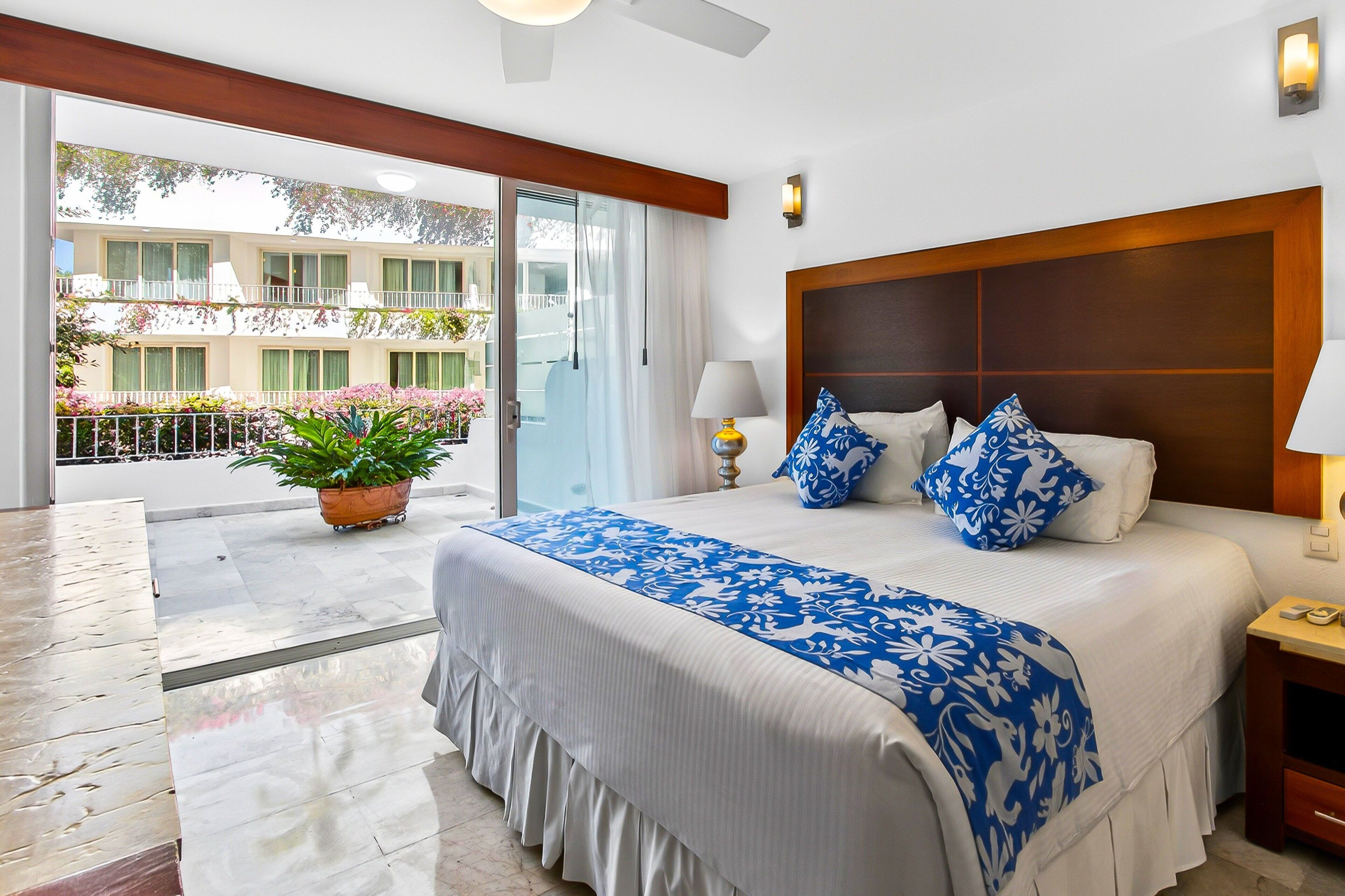 Habitación de hotel con una cama grande, un balcón con plantas en macetas, un cabecero de madera y una mesita de noche con lámpara.