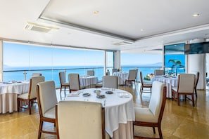 A dining area with round tables, chairs, and a large window overlooking the sea. - Well-located Hillside Suite With Rooftop Pool, Fitness Center & Family Features (Puerto Vallarta)