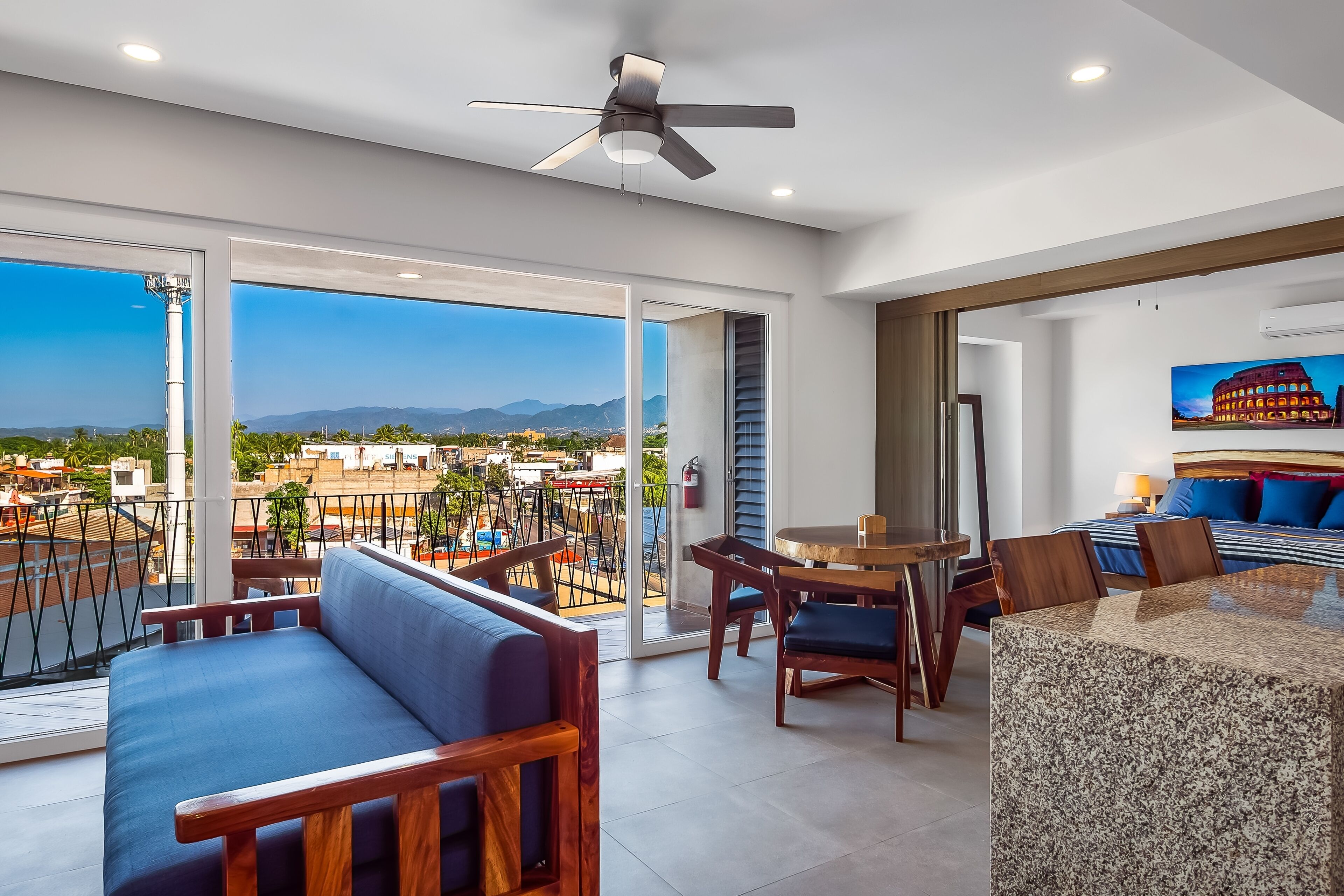 A modern hotel room with a balcony, a dining area, and a large window offering a city view.