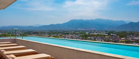 A rooftop pool with lounge chairs and a view of mountains and a city.