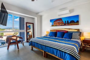 A bedroom with a large bed, a TV, a painting of the Colosseum, and a view of the city. - Resort Condo With Balcony, Views, Rooftop Pool, Gym, & Full Kitchen (Puerto Vallarta)