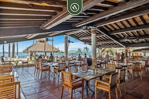 Ein Restaurant im Freien mit Holztischen und -stühlen, einem Reetdach und Blick auf das Meer.