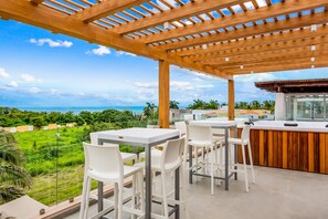 A covered outdoor seating area with tables and chairs, overlooking a lush green landscape and the ocean. - Ocean-view Getaway With Patio, Grill, Roof Deck, & On-site Pools (La Cruz de Huanacaxtle)