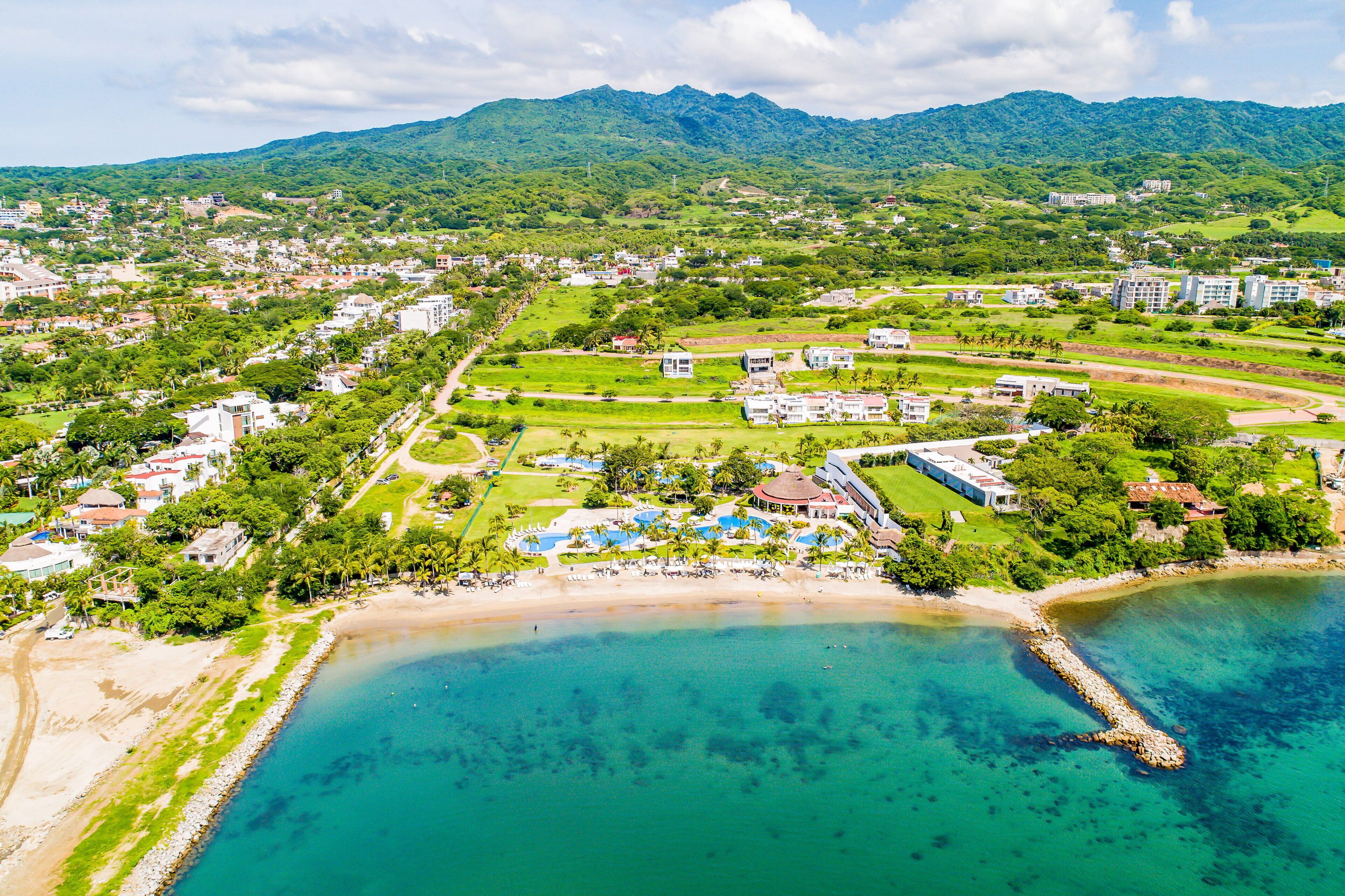 Vue aérienne d’une station balnéaire avec une plage, des piscines et un paysage montagneux en arrière-plan.