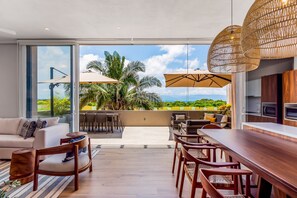 A modern living room with a large glass door leading to a balcony with outdoor furniture and a view of palm trees.