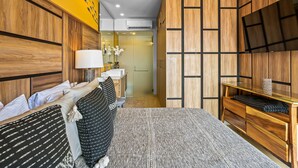 A modern hotel room with a wooden headboard, a bed with patterned bedding, a bedside table with a lamp, and a TV on a wooden stand. - Bright Coastal Condo With Pool Access, Private Balcony & Fast Wi-fi (Bucerías)