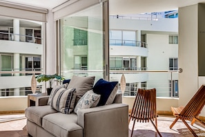Una sala de estar moderna con un sofá, una silla de madera y un ventanal con vistas a un edificio de varios niveles.