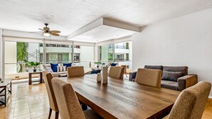 A spacious dining area with a large wooden table and comfortable chairs, large windows offering views of the outside, and a ceiling fan. - Nitta Condo # 104 Chic, Pool-view Condo With Furnished Balcony - Steps to Beach (Nuevo Nayarit)