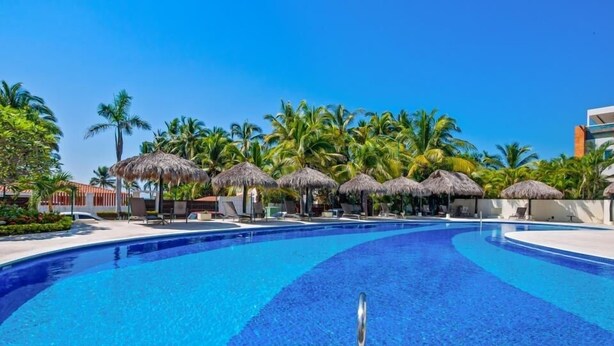 A swimming pool with a clear blue water, surrounded by palm trees and thatched umbrellas. - Nitta Condo # 104 Chic, Pool-view Condo With Furnished Balcony - Steps to Beach (Nuevo Nayarit)