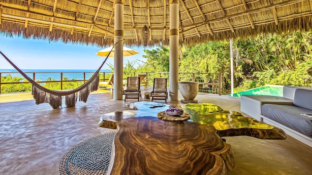 Zona de estar al aire libre con techo de paja y vistas al océano, con una hamaca y sillones junto a la piscina.
