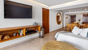Ein modernes Wohnzimmer mit Flachbildfernseher, einer Holzregal mit Büchern und Dekoration, einem Korbsessel und einem Bett mit weißer Bettwäsche.