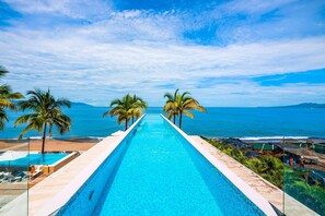 A long swimming pool with clear blue water, surrounded by palm trees and a view of the ocean. - Luxury Apartment With Infinity Pool, Ocean Views, Balcony & Easy Downtown Access (Puerto Vallarta)