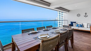 Ein Essbereich mit einem Tisch, gedeckt für sechs Personen, mit Blick auf ein Gewässer und Boote, ein Balkon mit Geländer und eine Couch mit gemusterten Kissen.