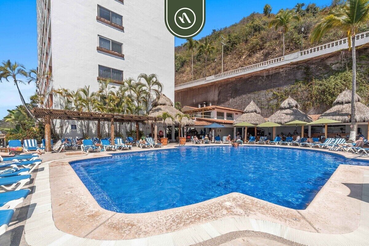 Uma área de piscina com espreguiçadeiras, um edifício com vários andares e uma encosta com estruturas de telhado de palha.