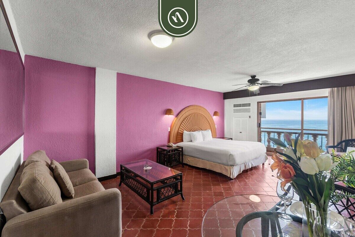 Habitación de hotel con cama, sofá, una mesita y balcón con vista al mar.