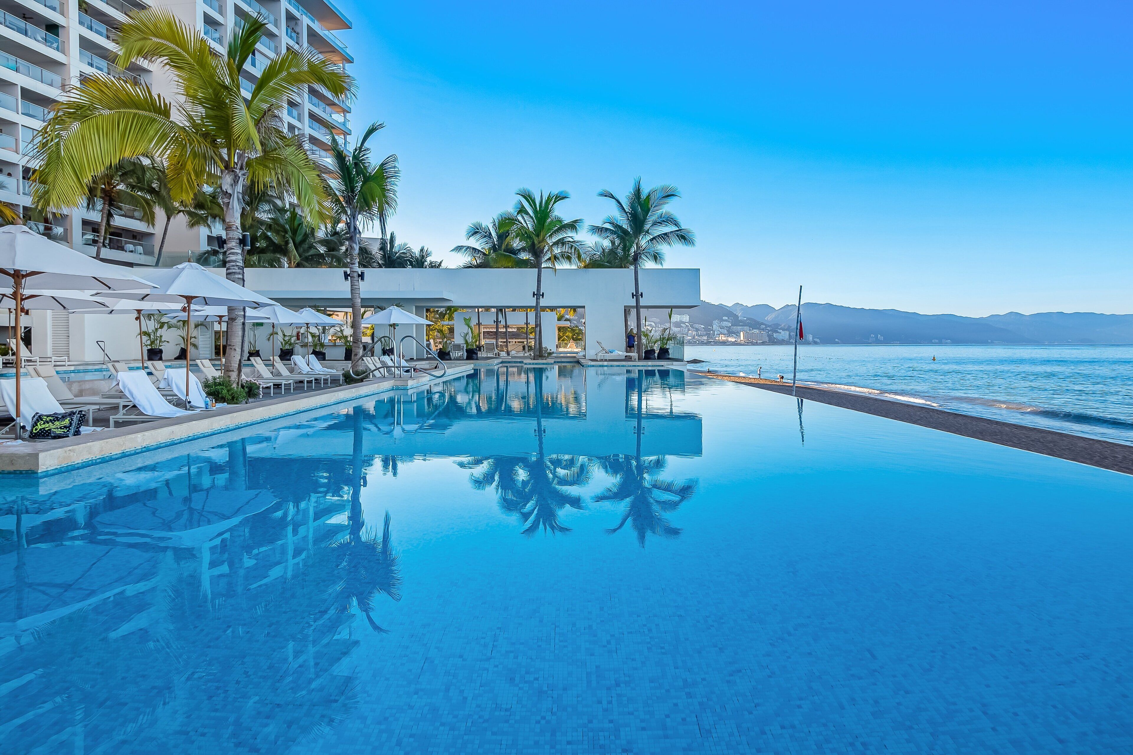 A poolside area with lounge chairs and umbrellas, a clear view of the ocean, and a modern building in the background.