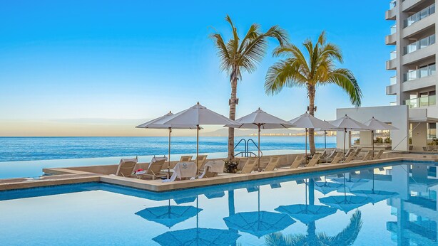 A poolside area with lounge chairs, umbrellas, and palm trees, overlooking a clear blue ocean. - Oceanfront Studio With Infinity Pool, Breathtaking Views & Beach Access (Puerto Vallarta)