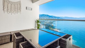 A balcony with a glass table and wicker furniture, overlooking a coastal city and ocean. - Oceanfront Studio With Infinity Pool, Breathtaking Views & Beach Access (Puerto Vallarta)