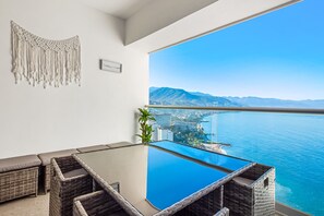 Ein Balkon mit Glastisch und Korbmöbeln, mit Blick auf eine Küstenstadt und das Meer.
