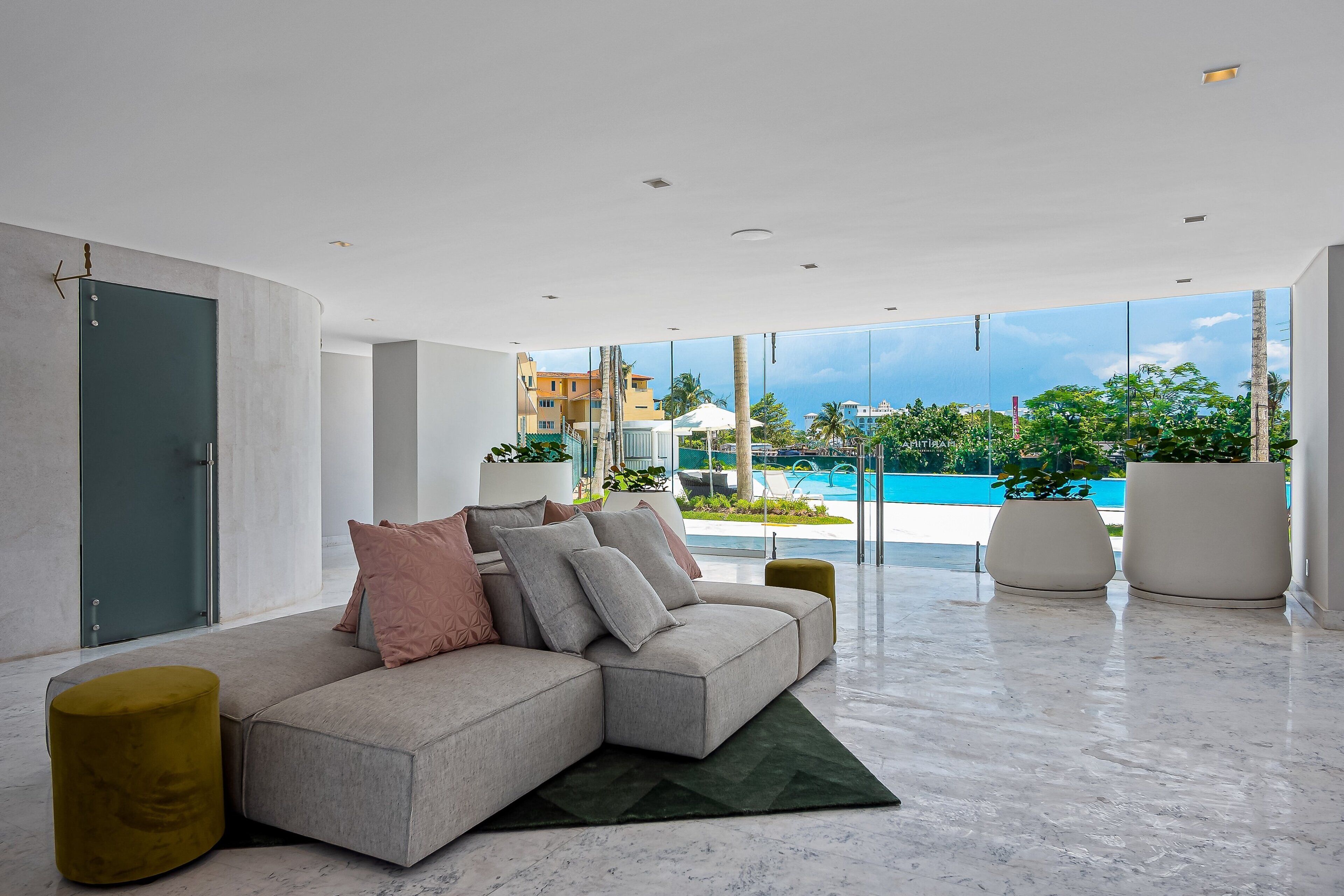 Un salon moderne avec un canapé d’angle, un tapis vert et une vue sur une piscine et des immeubles.