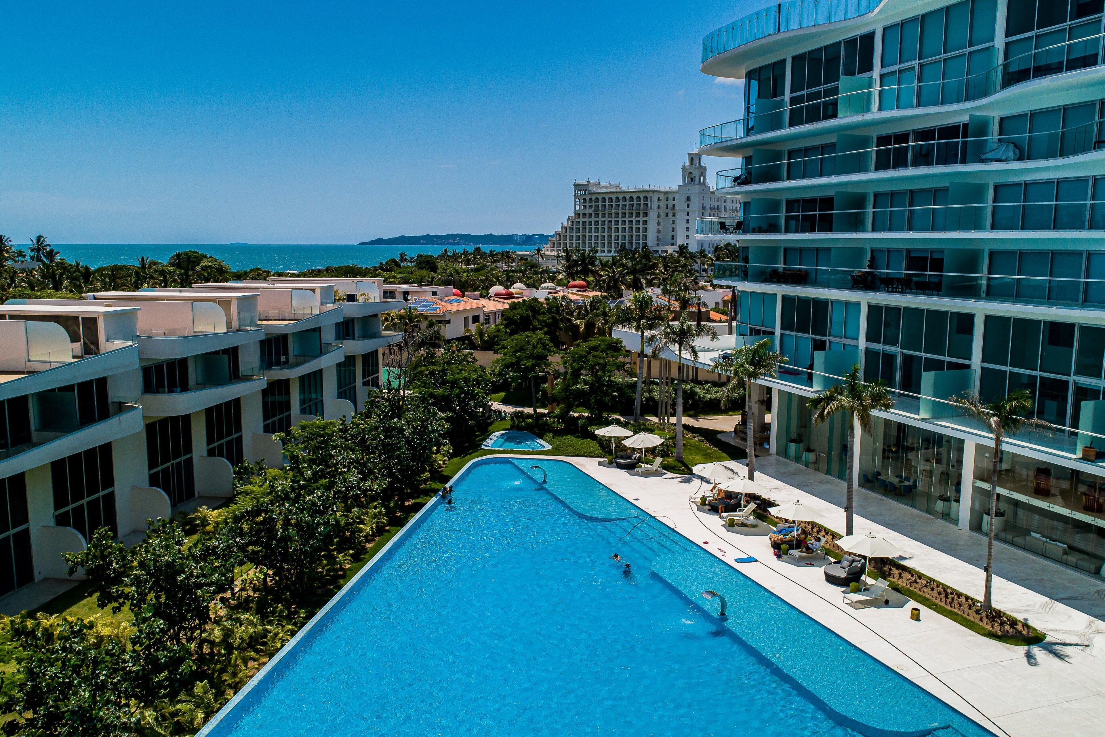 A resort with a large swimming pool, modern buildings, and a view of the ocean.