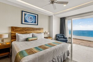 A hotel room with a large bed, a wooden headboard, a desk with a lamp, a chair, and a view of the ocean through a large window. - Modern Condo With Ocean Views, Rooftop Infinity Pool & Central AC (Puerto Vallarta)