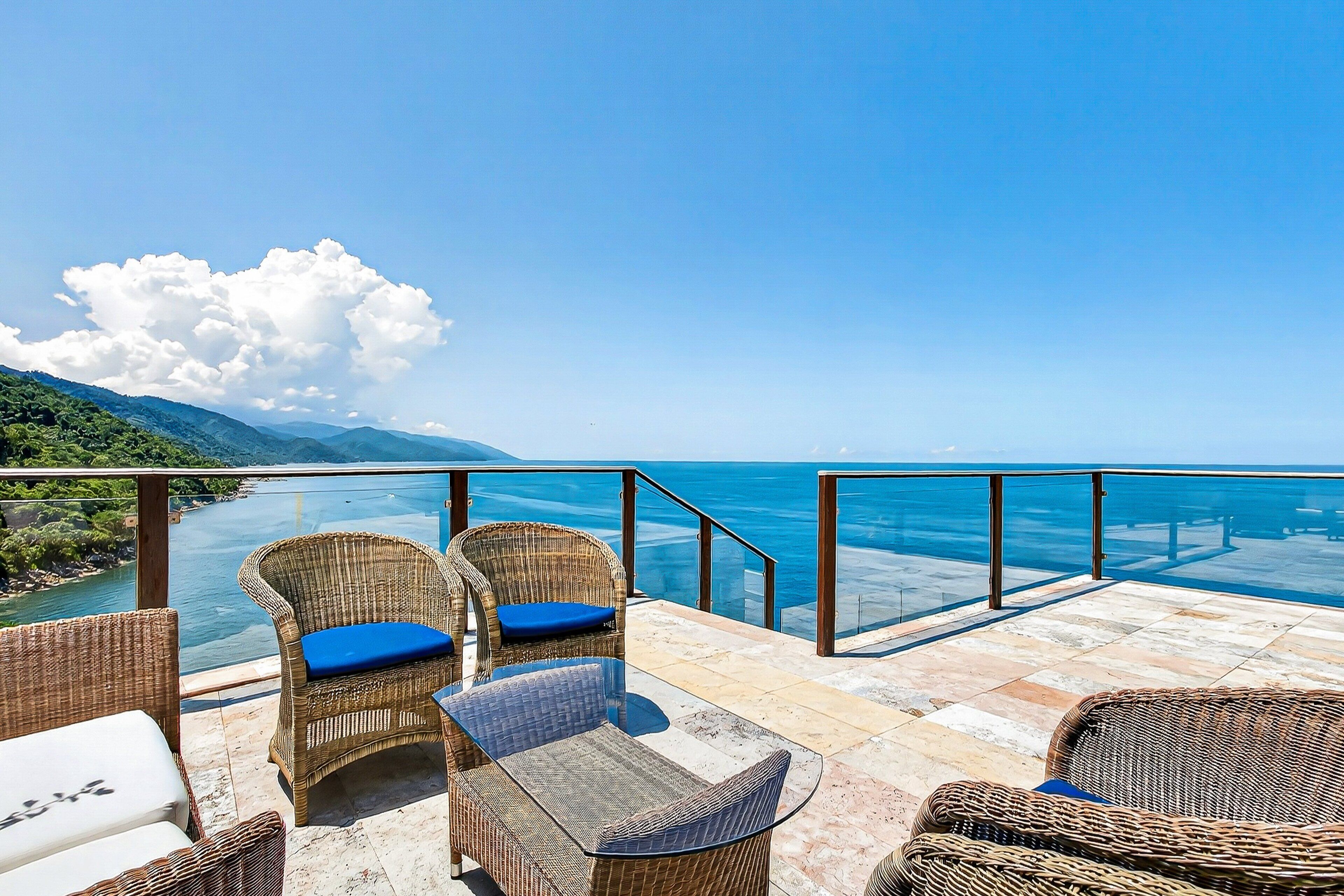 A terrace with wicker furniture, a glass table, and a view of the sea and mountains.