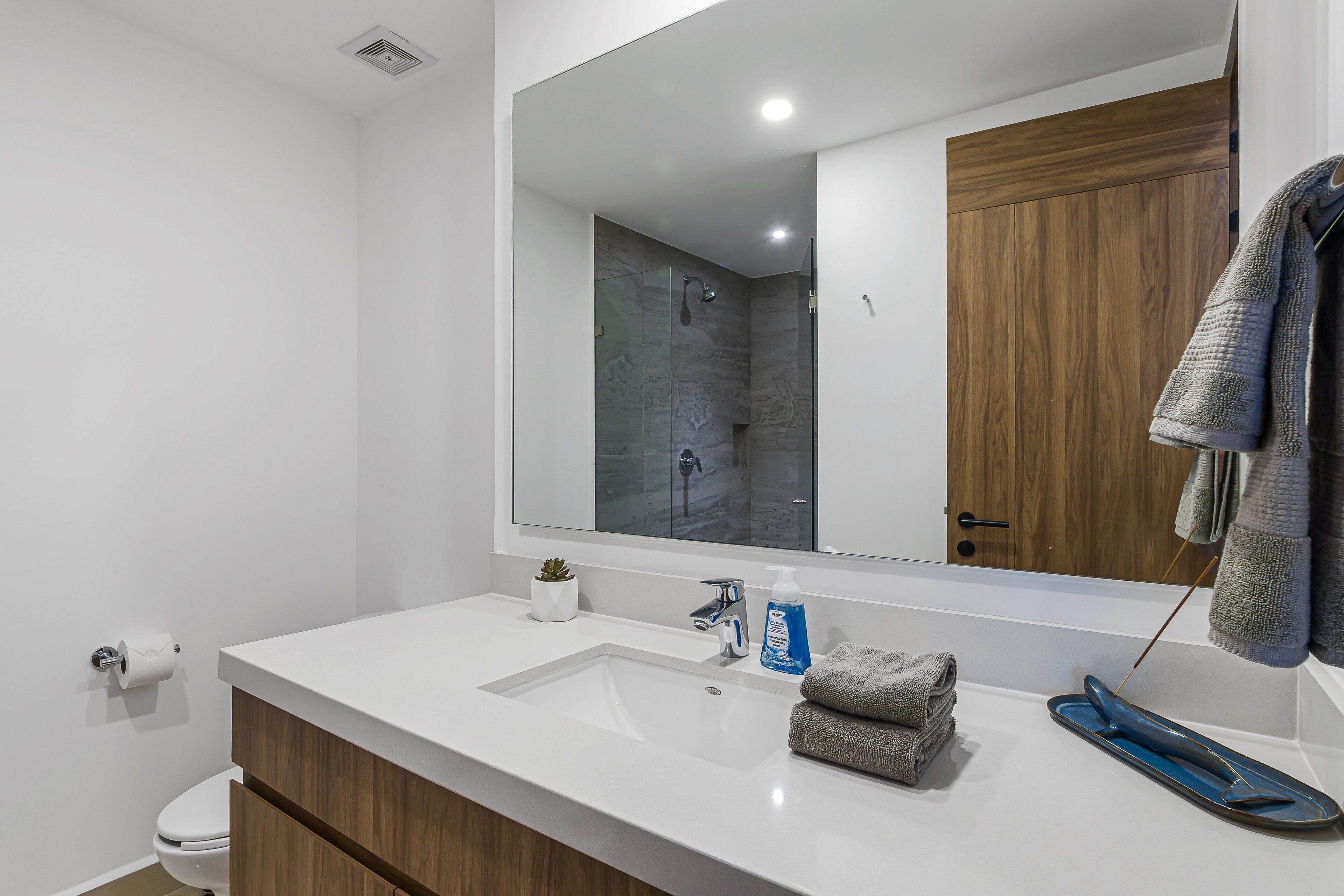 A modern bathroom with a white sink, wooden cabinet, and a large mirror reflecting a shower area.
