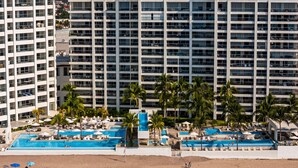A high-rise hotel with multiple balconies, a pool area with palm trees, and a sandy beach. - Stylish Oceanfront Condo With Infinity Pool, Rooftop Deck & Beach Access (Puerto Vallarta)