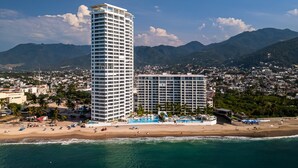 A high-rise hotel with a pool and beach access, surrounded by mountains and a coastal town. - Modern Oceanfront Studio With Breathtaking Sunsets, Heated Pools & Beach Access (Puerto Vallarta)
