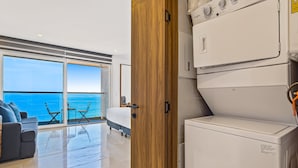 A modern hotel room with a large sliding glass door leading to a balcony with a view of the sea, a sofa, a small table, and a washing machine. - Modern Oceanfront Studio With Breathtaking Sunsets, Heated Pools & Beach Access (Puerto Vallarta)
