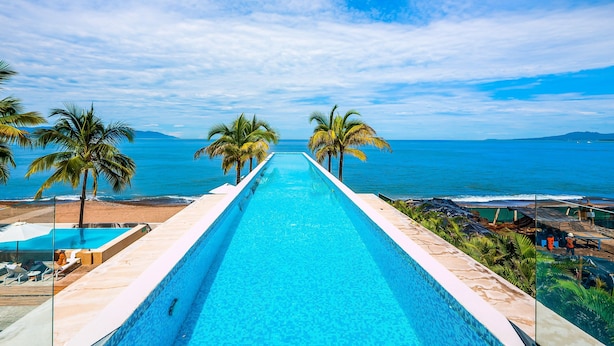 A long swimming pool with a view of the ocean and palm trees. - Modern Oceanfront Studio With Breathtaking Sunsets, Heated Pools & Beach Access (Puerto Vallarta)