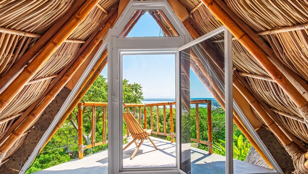 A triangular room with a wooden chair on a balcony, a thatched roof, and a view of greenery and the sea. - Relaxing Apartment With Mini Pool & High-speed Internet - Explore Local Delights (La Culebra)