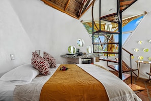 A bedroom with a bed, pillows, a desk, a chair, and a view of the outdoors.