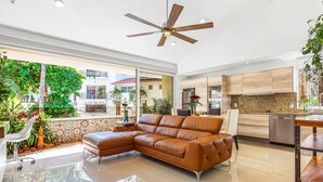 Un soggiorno moderno con un ampio divano in pelle marrone, una ventola a soffitto e un angolo cottura con mobili in legno e un frigorifero in acciaio inossidabile.