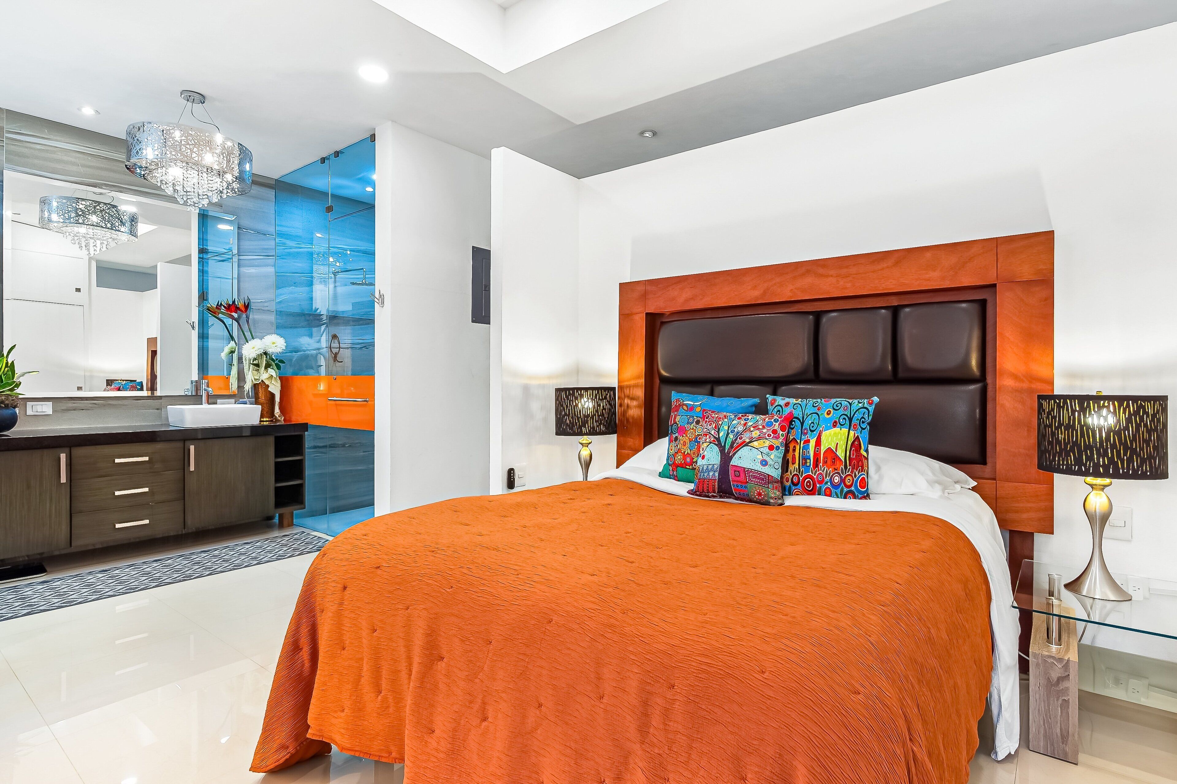 A modern bedroom with a large bed, a nightstand, a lamp, and a bathroom visible in the background.