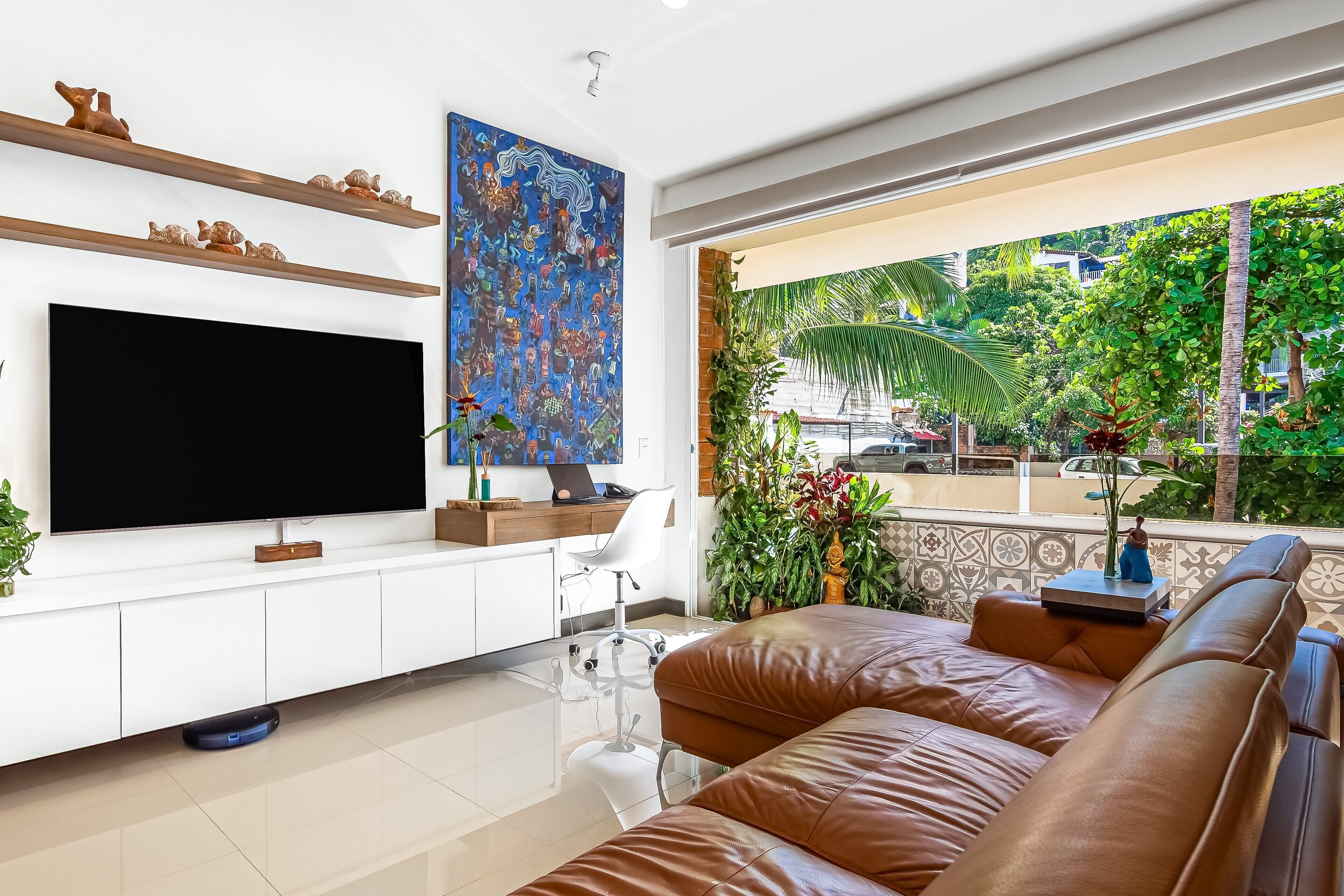 Ein modernes Wohnzimmer mit großem Flachbildfernseher, einem braunen Ledersofa, einem weißen Schreibtisch mit Computer und einem großen Fenster mit Blick auf Grünflächen und Gebäude.