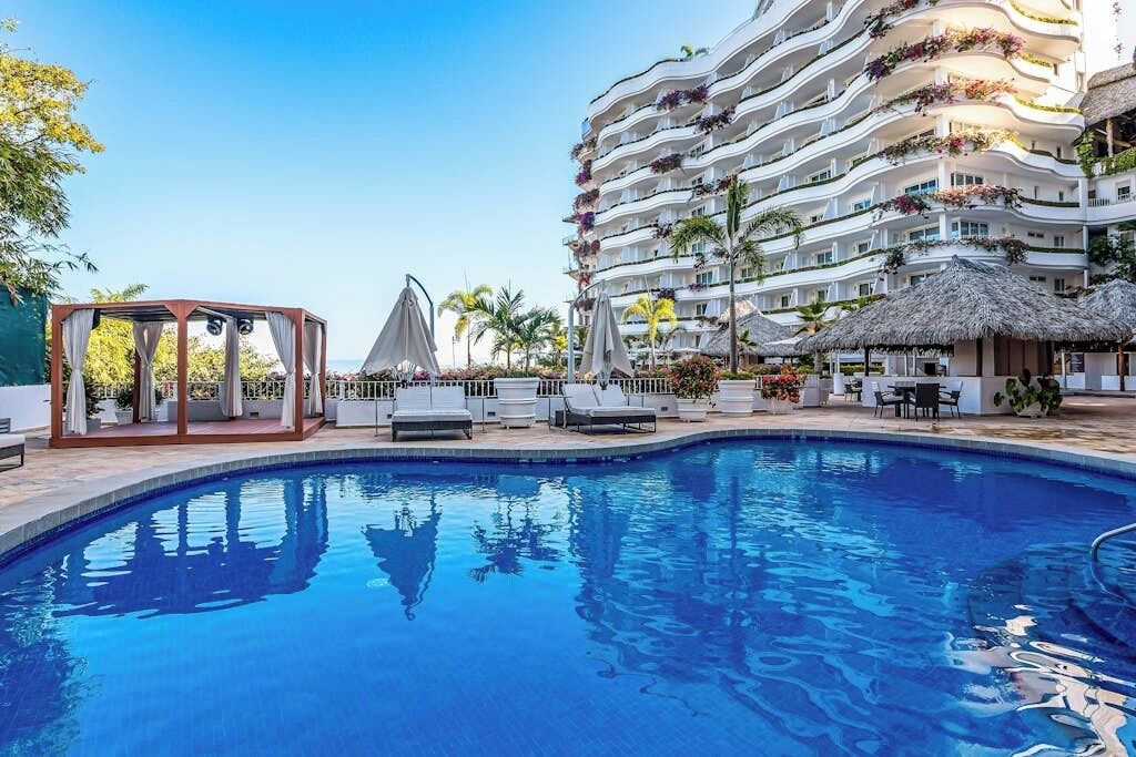 Una piscina con sillas de descanso y un pabellón, rodeada por un hotel de varios pisos con balcones.