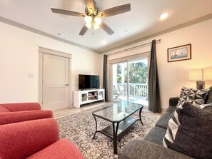 Ein Wohnzimmer mit Deckenventilator, einem Fernseher auf einem weißen Schrank, einem Glastisch, roten und grauen Sofas und Blick durch eine Glastür zu einer Terrassenanlage.