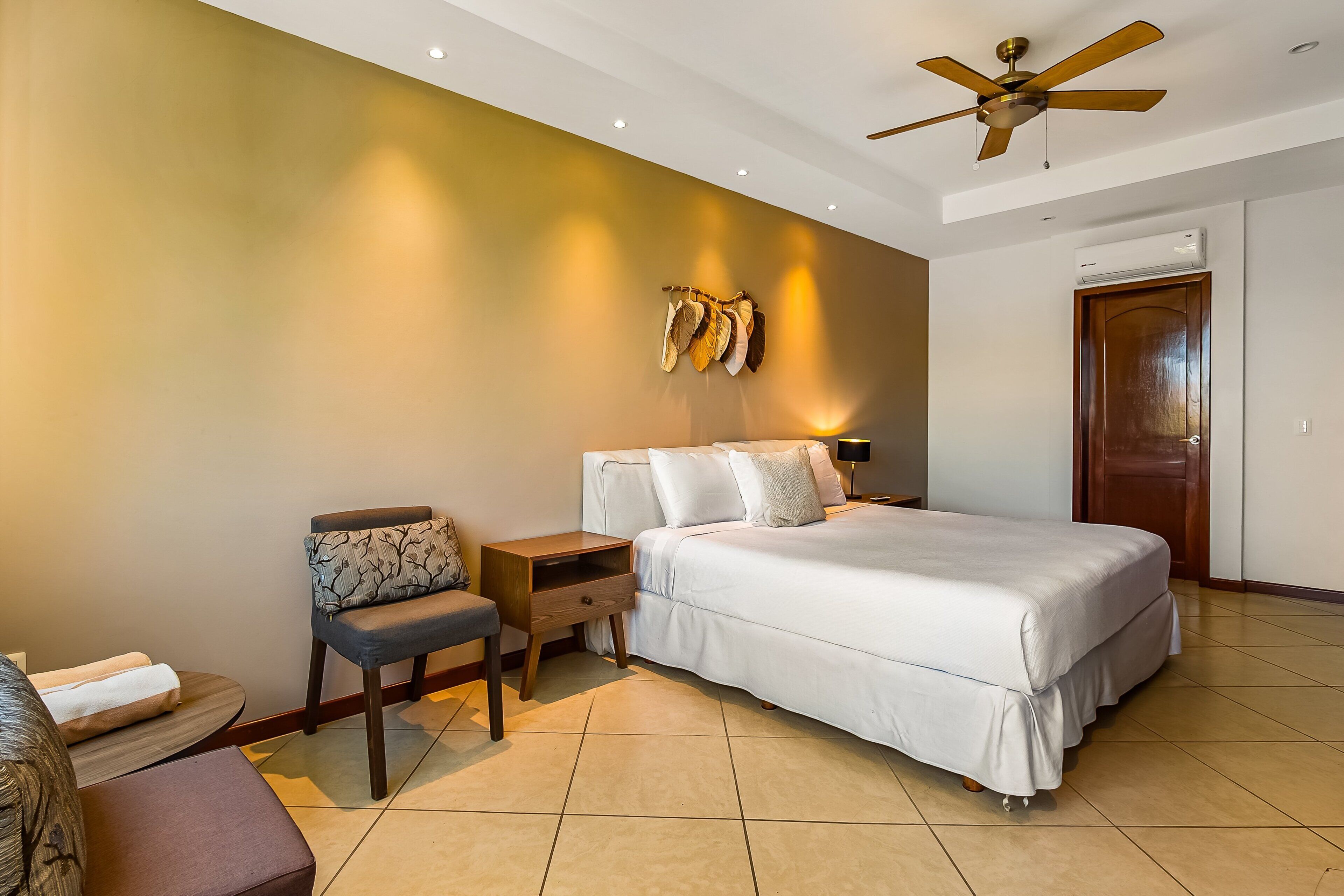 Une chambre à coucher comprenant un lit, des tables de chevet, une chaise, un ventilateur de plafond et un appareil de climatisation.
