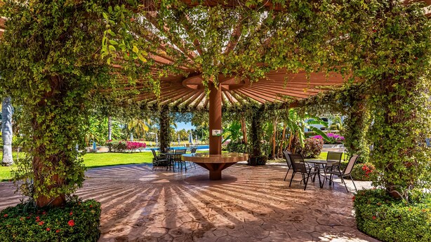 A covered outdoor seating area with a central table and chairs, surrounded by greenery and a paved walkway. - Cozy Condo With Loft, Private Balcony and Gorgeous Shared Pool (Nuevo Nayarit)