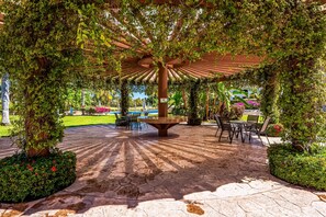 A covered outdoor seating area with a central table and chairs, surrounded by greenery and a paved walkway. - Cozy Condo With Loft, Private Balcony and Gorgeous Shared Pool (Nuevo Nayarit)