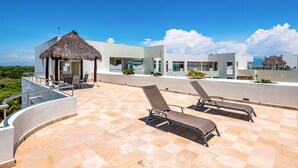 A rooftop terrace with lounge chairs, a thatched-roof structure, and modern buildings in the background. - Ocean-view Condo With Resort Pool & Sundeck (Nuevo Nayarit)