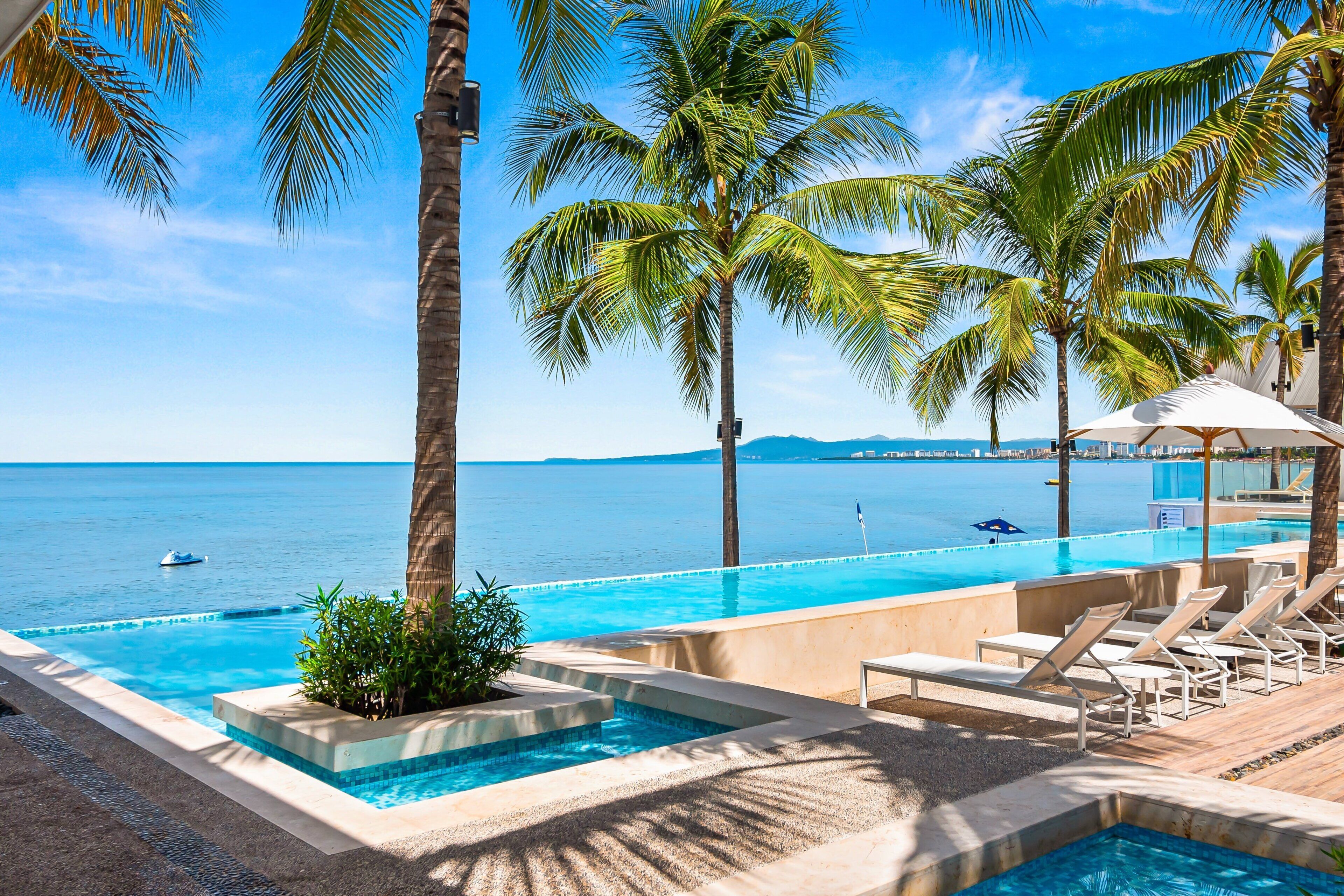 A pool area with lounge chairs, palm trees, and a view of the sea.
